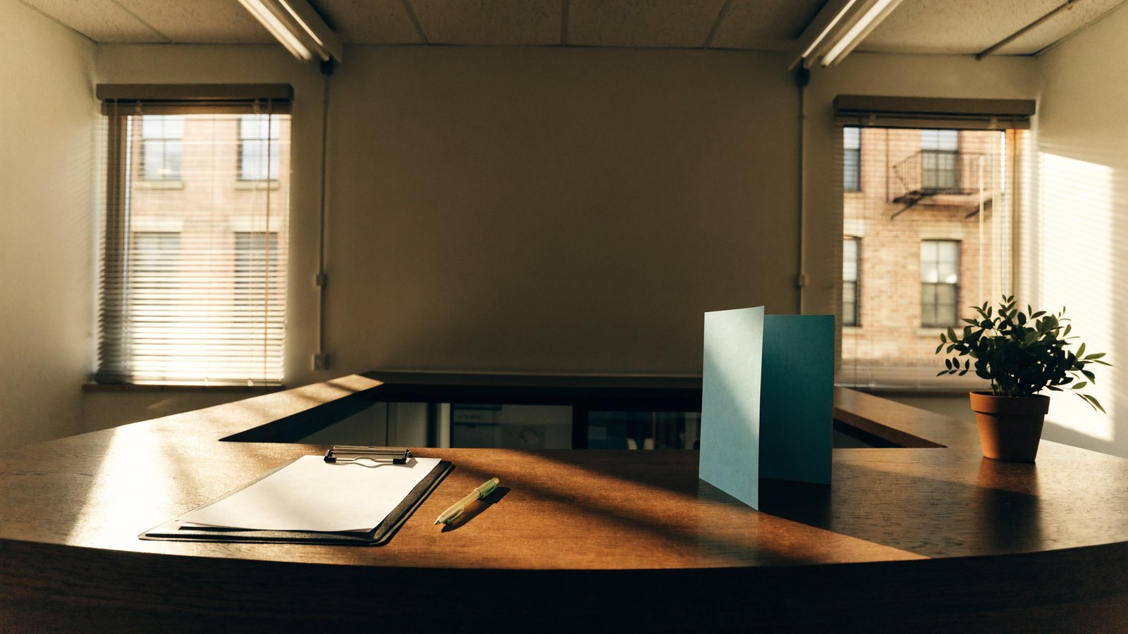 A small hospital front desk in warm light with a clipboard, a pen, a plain teal brochure and a potted plant.