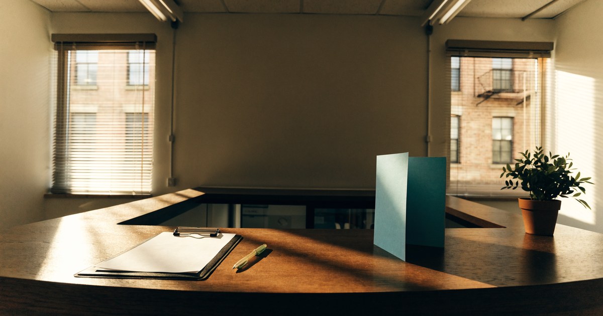A small hospital front desk in warm light with a clipboard, a pen, a plain teal brochure and a potted plant.